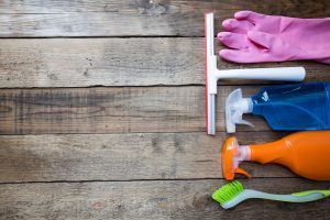 Various cleaning supplies including pink gloves, squeegee, and spray bottles on a wooden background for blog page